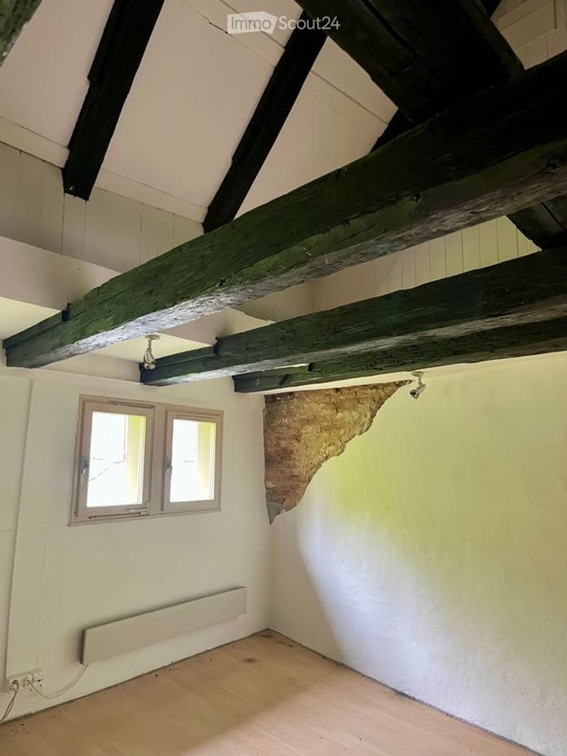 Empty room with exposed wooden beams on the ceiling, wooden flooring, two windows, and a radiator on one wall.