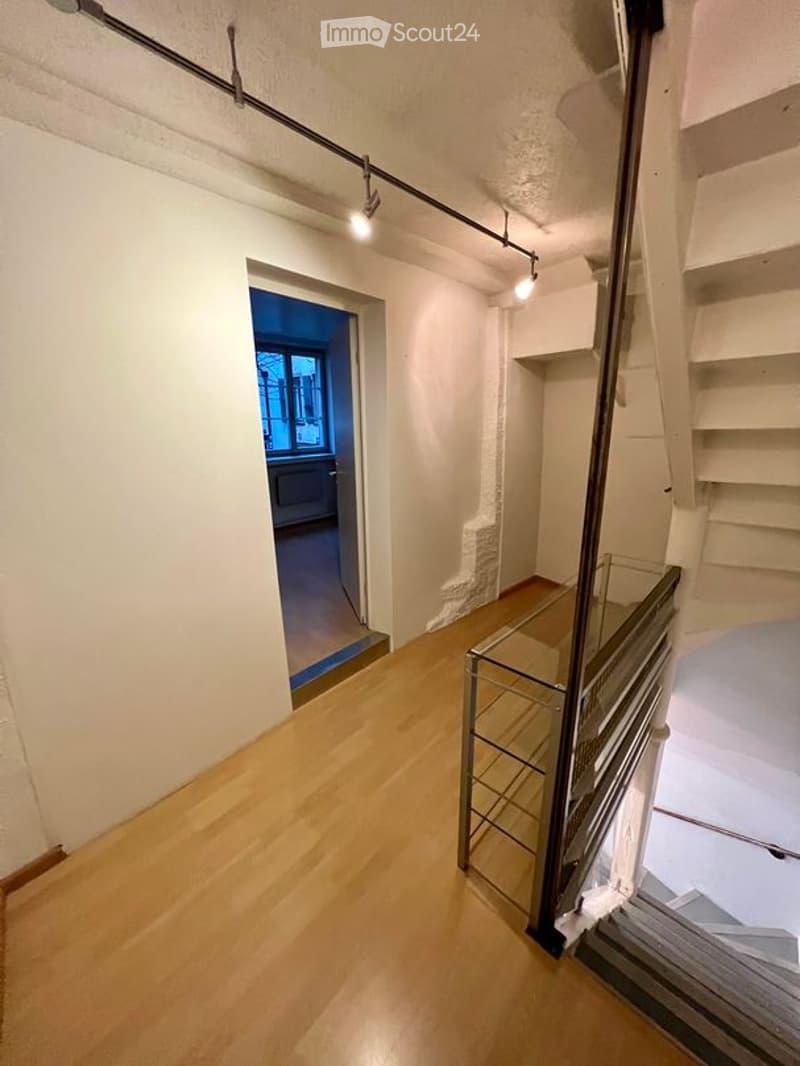 A narrow hallway with light wooden flooring, white walls, and a glass railing leading to a staircase. A small room with a window is visible at the end of the hallway.