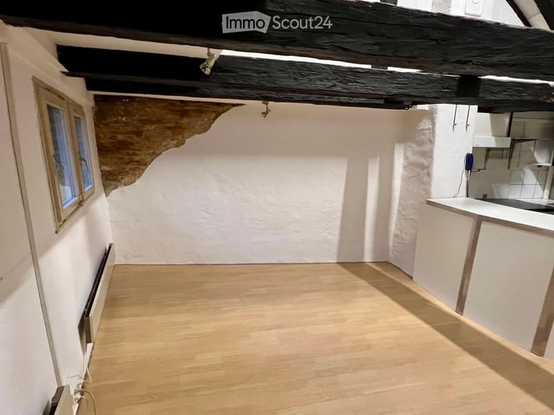 Empty room with wooden flooring, white walls, exposed wooden beams on the ceiling, and a small window on the left side. The room has a small kitchen area with white cabinets and a countertop on the right side.
