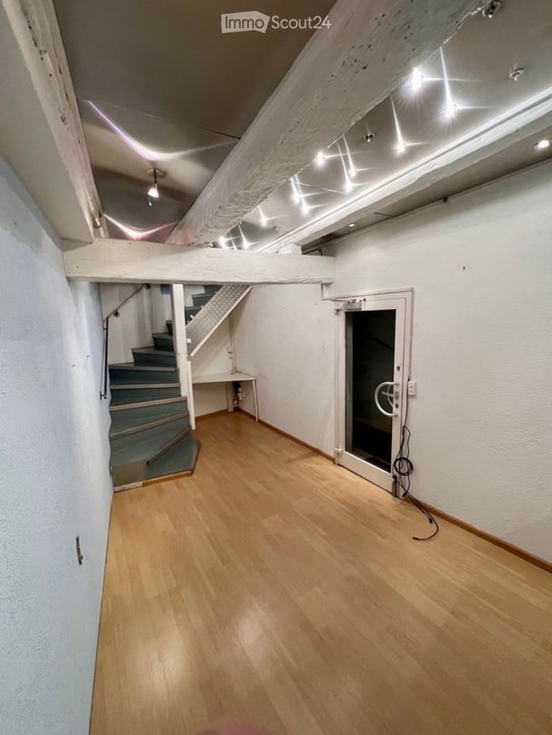 Empty room with wooden flooring, white walls, and a staircase leading to an upper level. The space includes a small table and a door leading to another area.