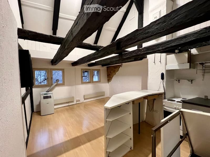 Bright kitchen with wooden floors, exposed wooden beams, white cabinets, and a central counter. Features include an oven, shelves, and an air conditioner unit.