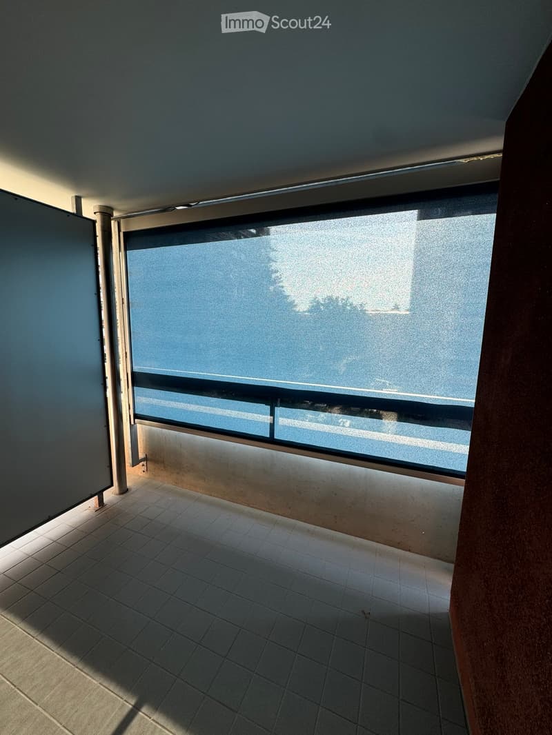 Empty balcony with tiled floor, large window, and metal railing, allowing natural light and a view of the outside.