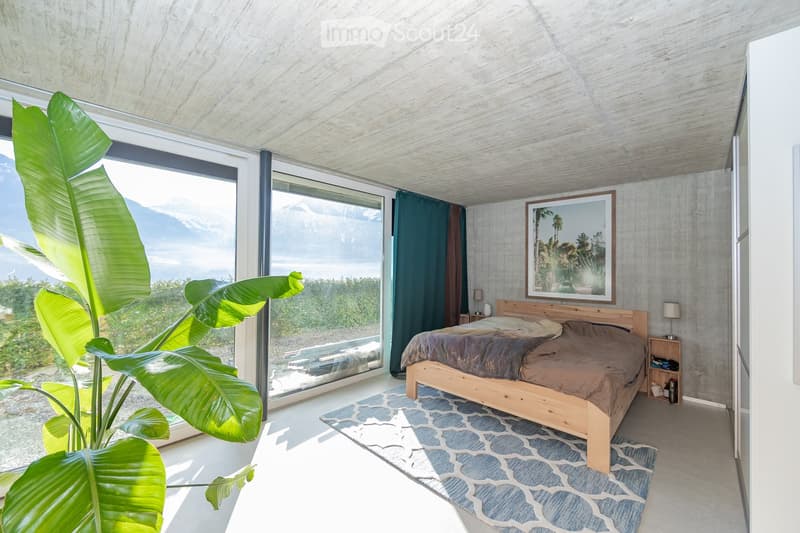 Modern bedroom with large windows offering a scenic mountain view, wooden bed, nightstands, and a large leafy plant near the window.