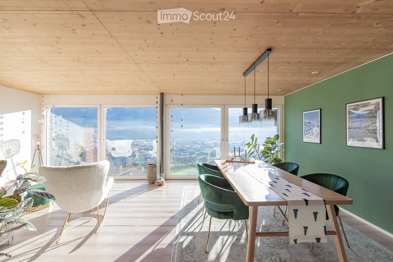 Modern dining area with a long wooden table, green velvet chairs, pendant lights, and large windows offering a scenic view.