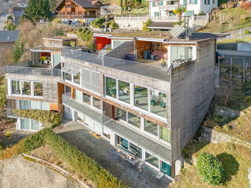 Modern two-story house with large glass windows, wooden accents, and multiple balconies overlooking a scenic hillside with neighboring houses and greenery.