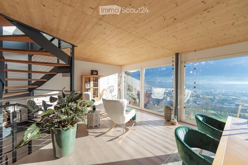 Modern living room with large windows offering a scenic mountain view, featuring a white sofa, green armchairs, a small table, a bookshelf, and a large potted plant.