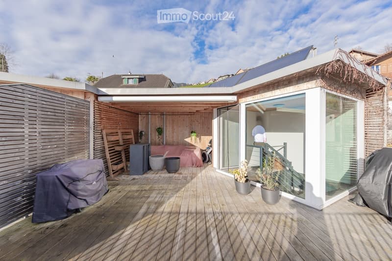 Wooden deck area with covered seating, potted plants, covered grill, and modern glass-walled structure.