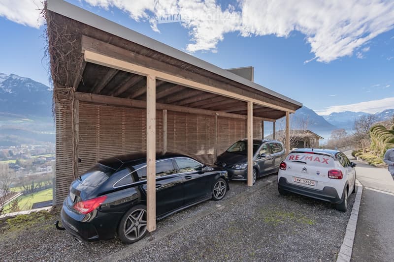 A modern wooden garage with a slanted roof, featuring three parked cars underneath. The background shows a scenic mountain view and a clear blue sky.