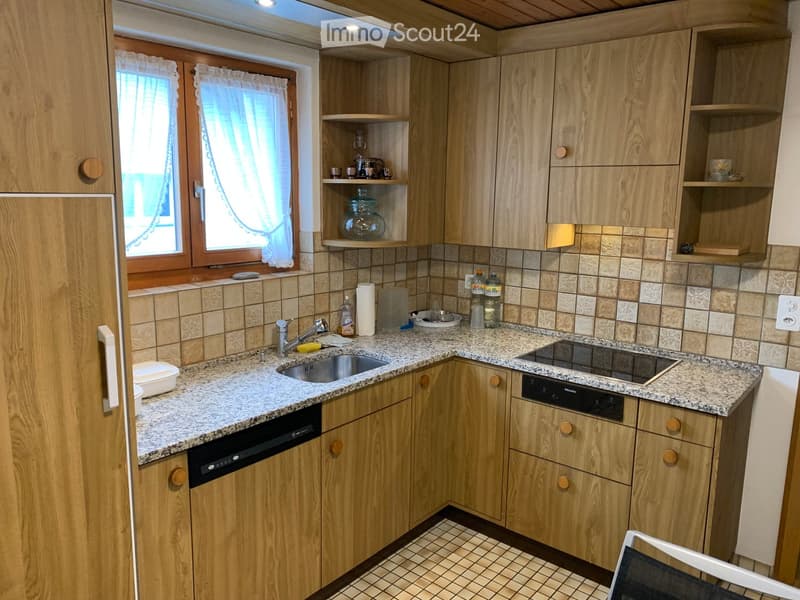 A modern kitchen with light wooden cabinets, granite countertops, a sink, a dishwasher, and a stove. The space features tiled backsplash and a window with white curtains.