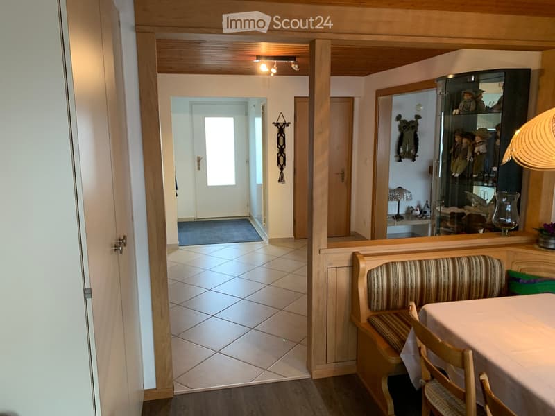 Bright hallway with tiled floor, wooden bench, dining table, chairs, and a mirror reflecting a display cabinet with dolls and decorative items.