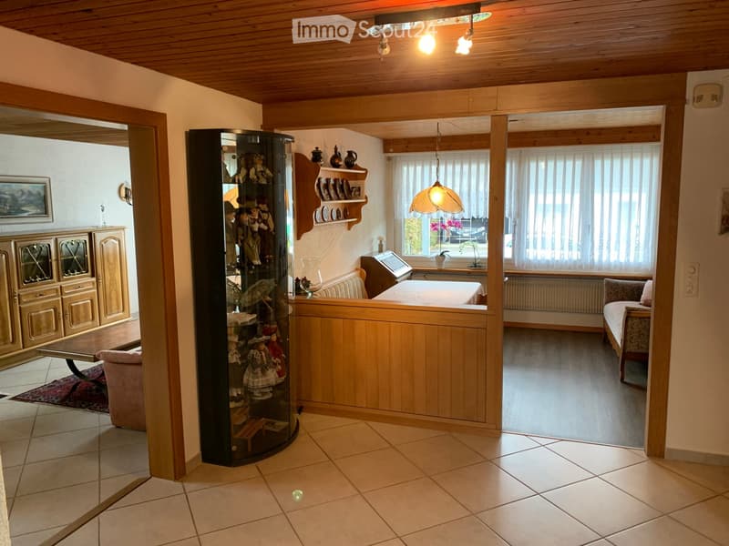 A well-lit living room with wooden beams, a sofa, a display case with dolls, shelves, a table, and a cabinet.