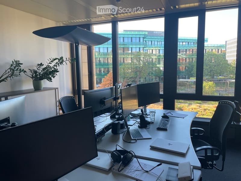 Modern office space with large windows, a desk with multiple monitors, a laptop, keyboard, mouse, headphones, and a potted plant on a shelf.