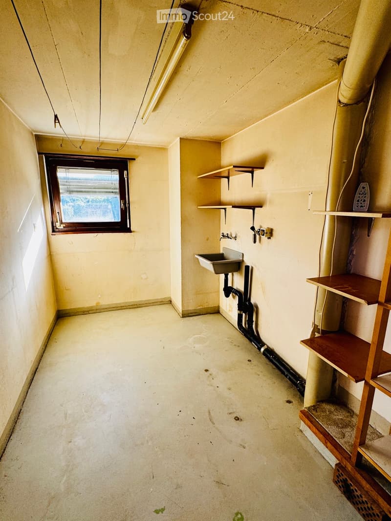 Empty room with concrete floor, exposed pipes, a small sink, shelves, and a window with a view of the outside.