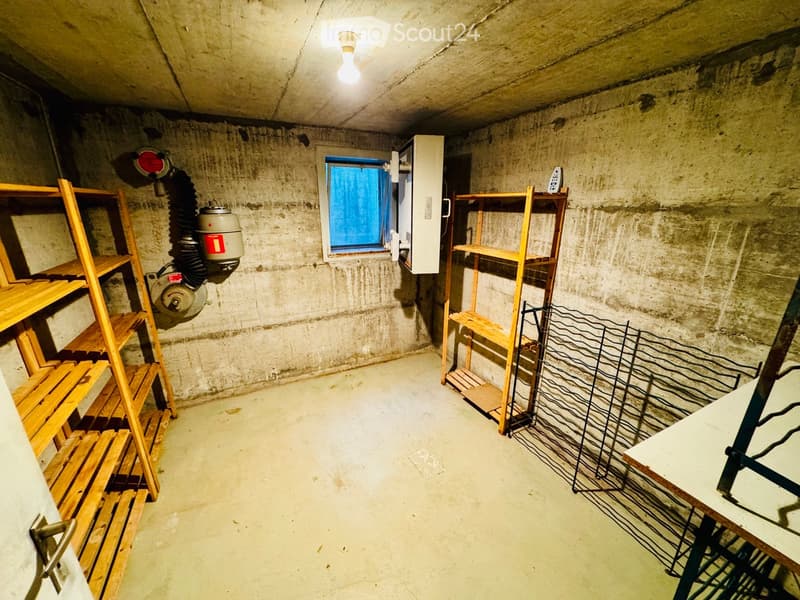 Empty basement room with concrete walls and floor, wooden shelving units, a small window with blue blinds, and a ventilation system.