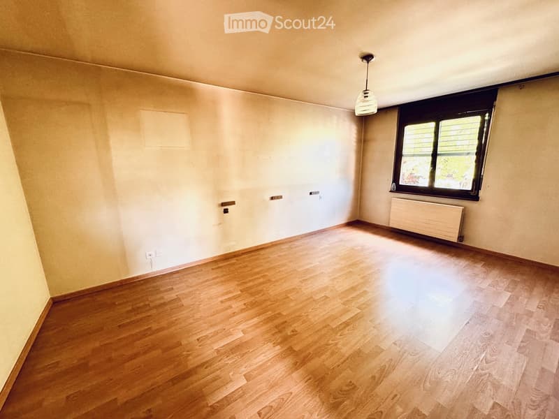 Empty room with wooden flooring, white walls, and a single window with blinds. A radiator is visible under the window, and a ceiling light fixture is present.