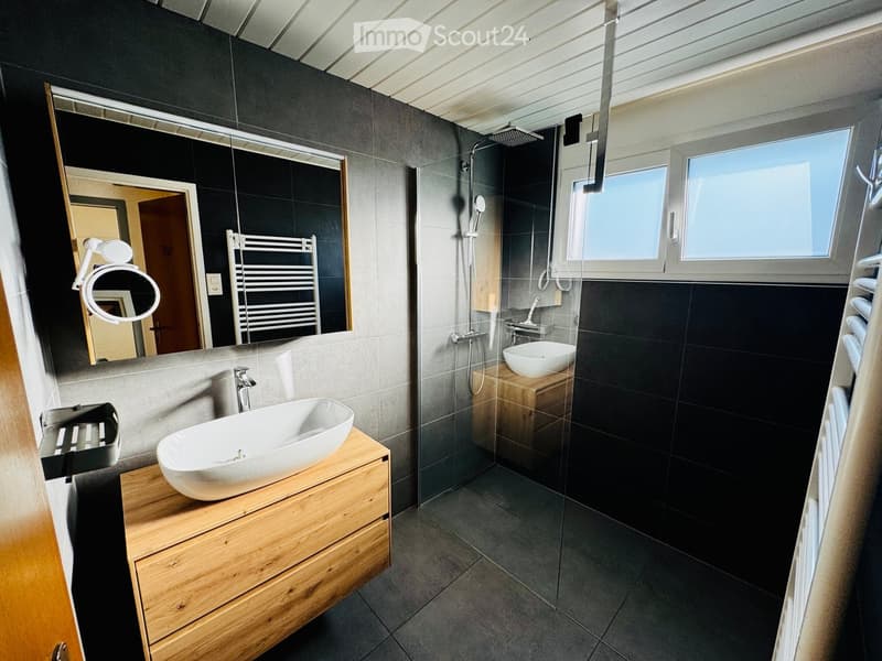 Modern bathroom with dark grey tiles, two sinks on wooden vanities, large mirror, glass-enclosed shower, and towel rack.