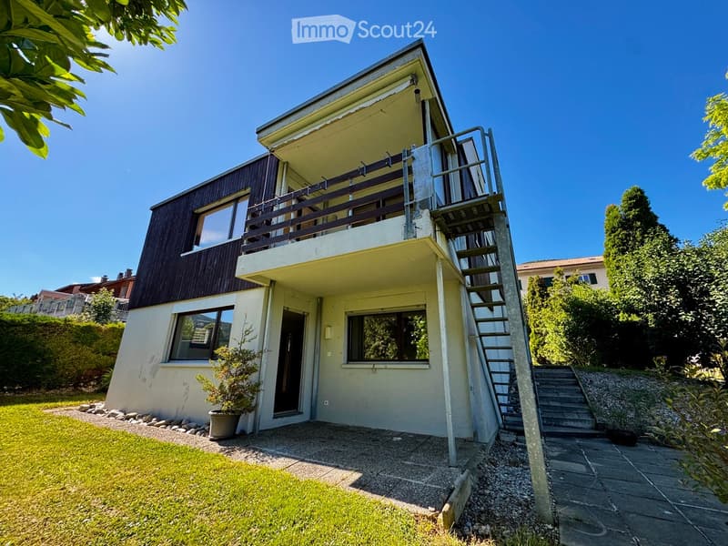Two-story house with a raised ground floor, balcony, and modern design. Features include a staircase leading to the upper level, a small garden with a potted plant, and a clear view of the surrounding greenery.