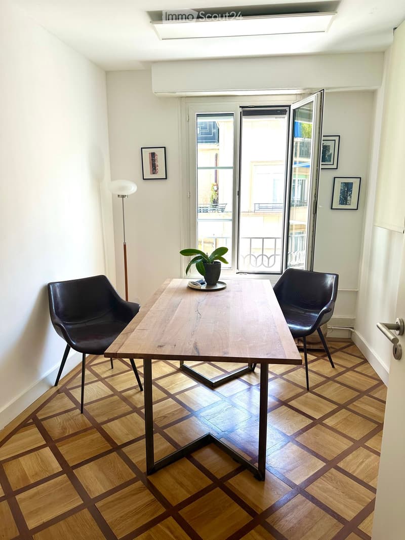 Bright room with wooden flooring, a long wooden table, two black leather chairs, a potted plant, a floor lamp, and framed artwork on the walls. Large sliding glass doors open to a balcony.