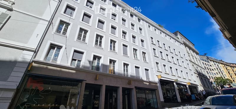 A multi-story building with white facade and numerous windows, featuring a storefront on the ground floor with glass windows and signage.