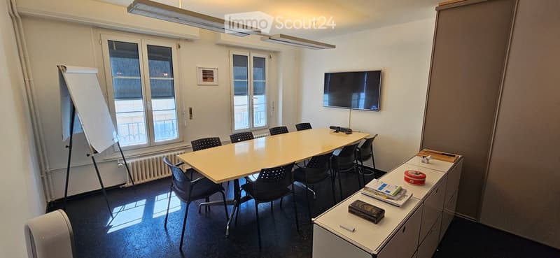 Bright meeting room with large windows, a long rectangular table surrounded by black chairs, a mounted screen, a cabinet with office supplies, and a whiteboard easel.