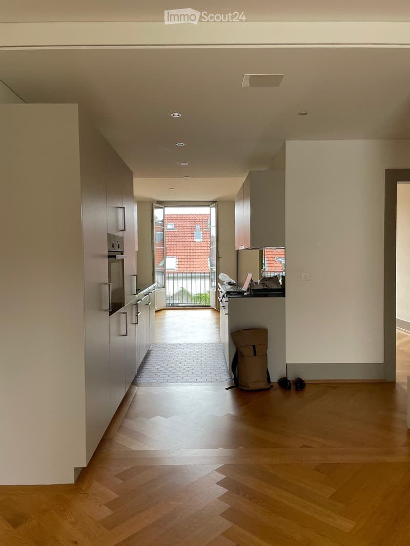 Modern kitchen with wooden flooring, white cabinets, stainless steel appliances, and a laptop on the counter. A backpack is placed near the entrance leading to a balcony.