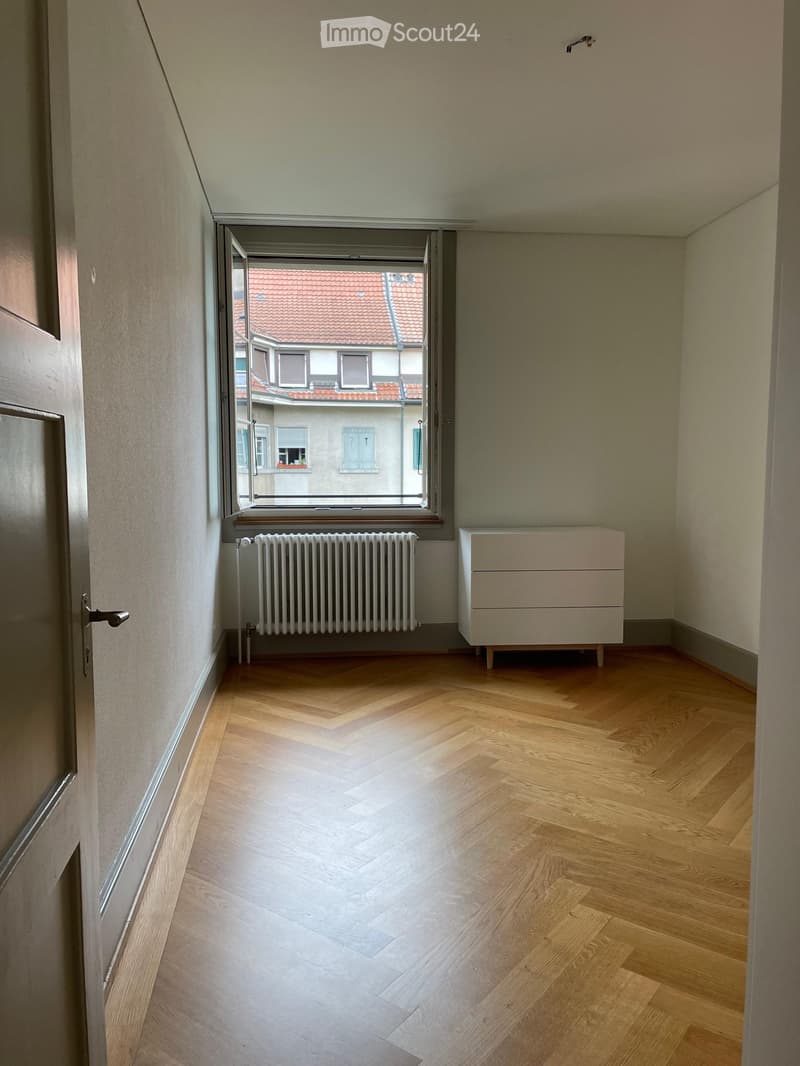 Empty room with wooden herringbone flooring, white walls, a radiator, and a small cabinet. A window offers a view of a neighboring building with a red-tiled roof.