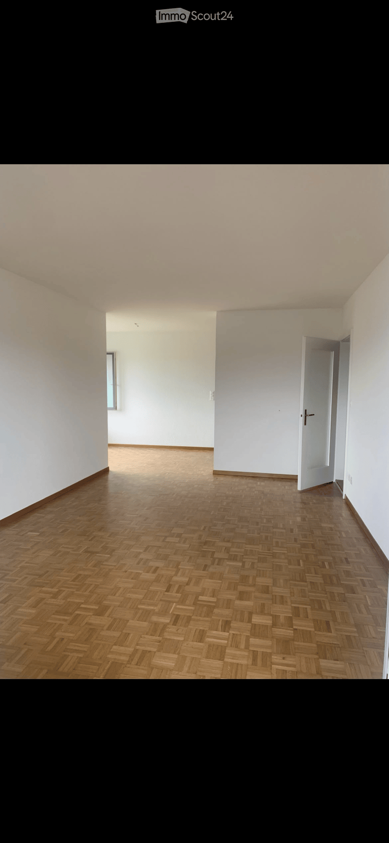 Empty room with wooden herringbone flooring, white walls, and a single window on one side. A door is visible on the right side of the room.