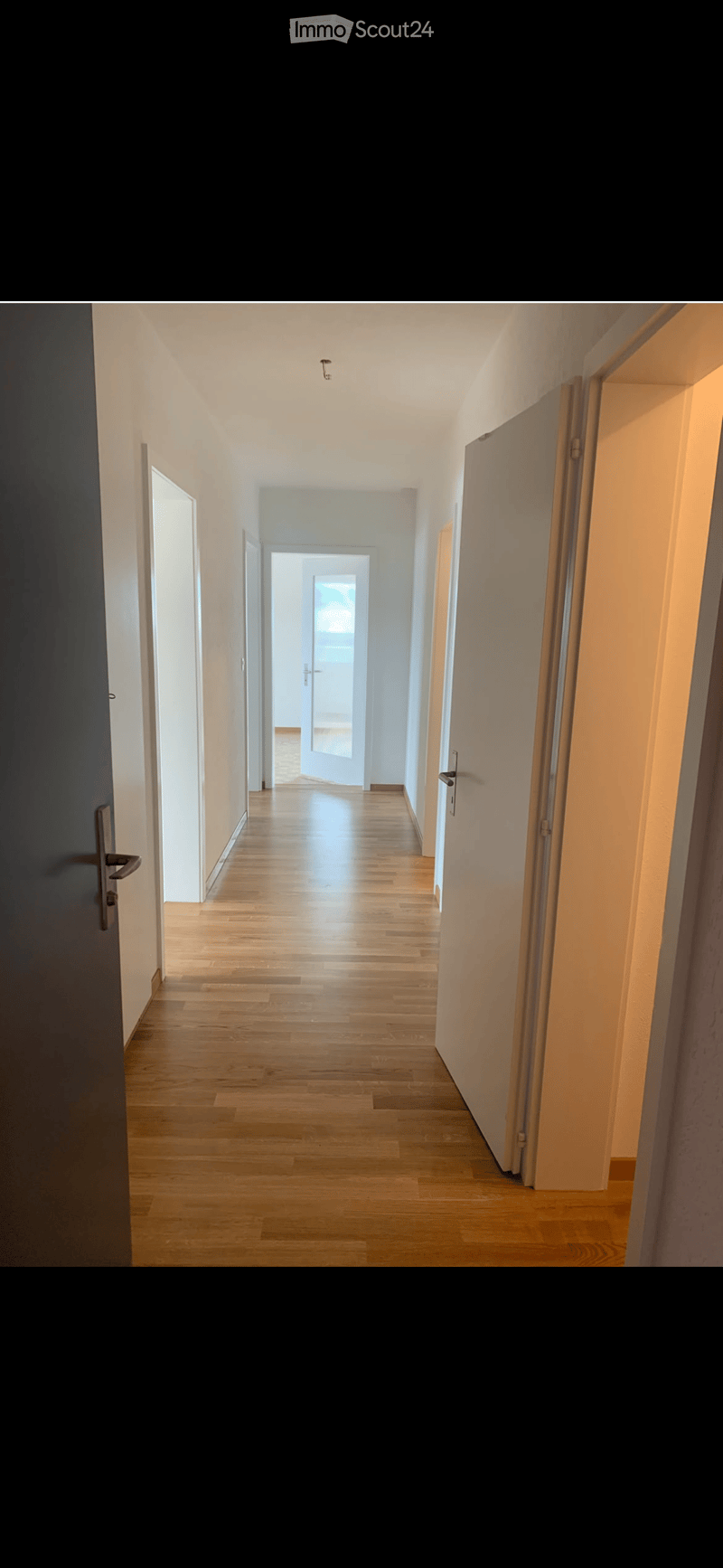 A long, narrow hallway with wooden flooring and white walls, featuring several doors on both sides and a bright light fixture in the ceiling.