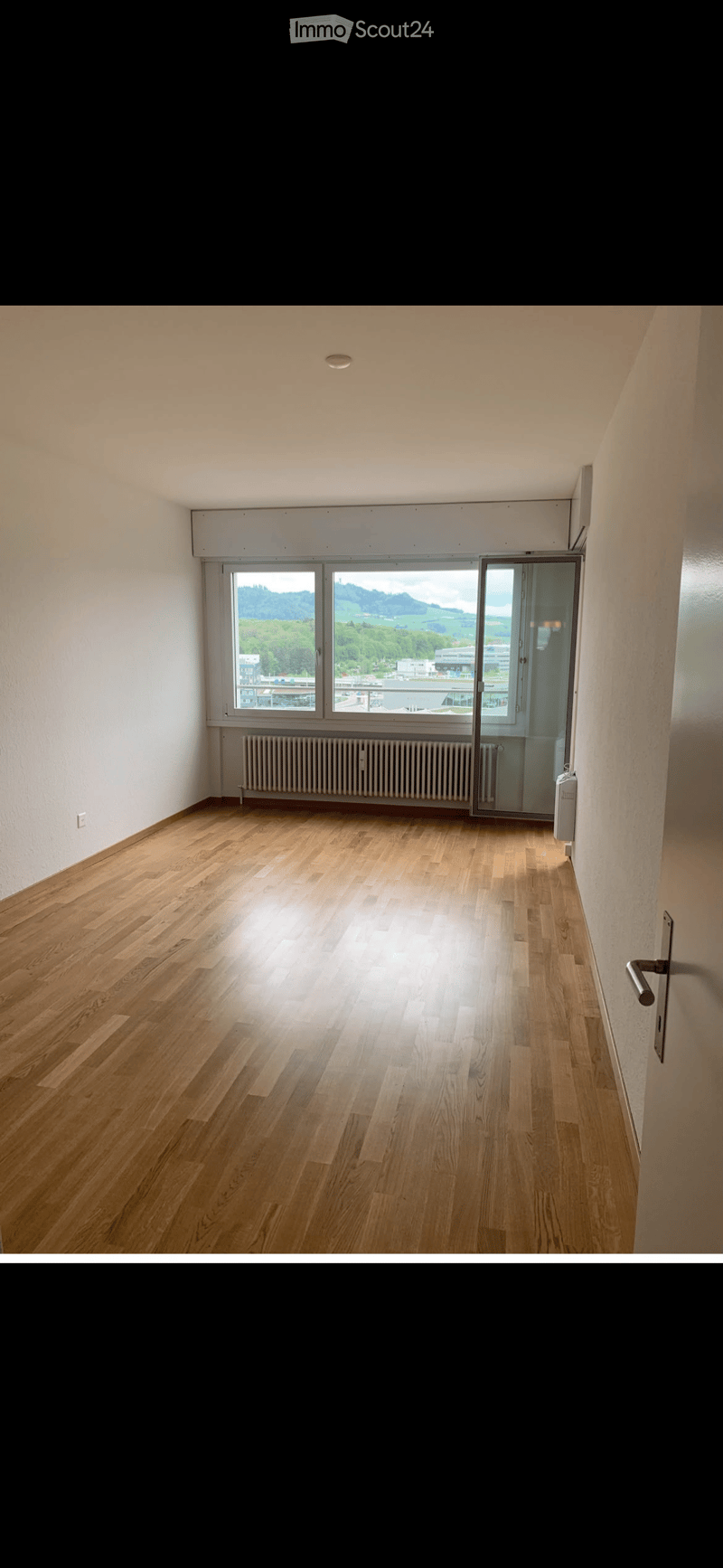Empty room with wooden flooring, white walls, large windows, and a radiator. No furniture or equipment present.