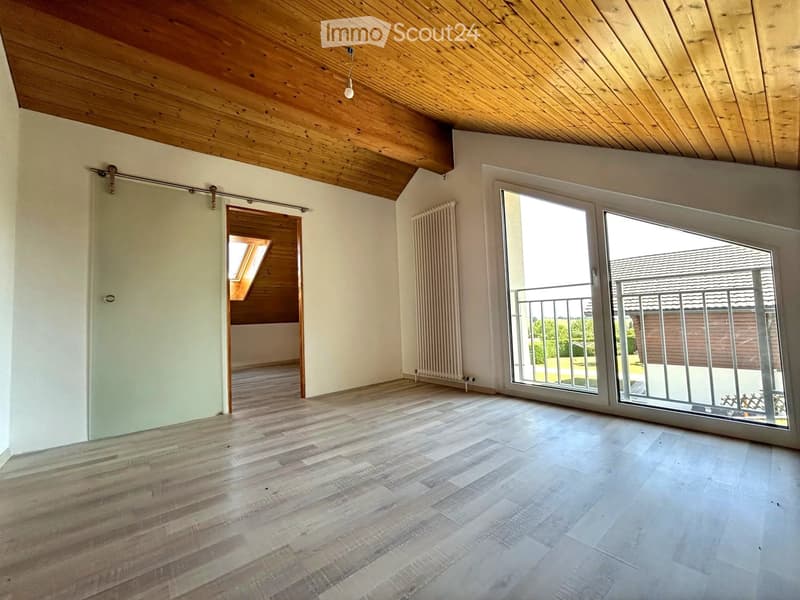 Bright, empty room with wooden flooring, white walls, and a large sliding glass door leading to a balcony. Features a wooden ceiling and a radiator on one wall.