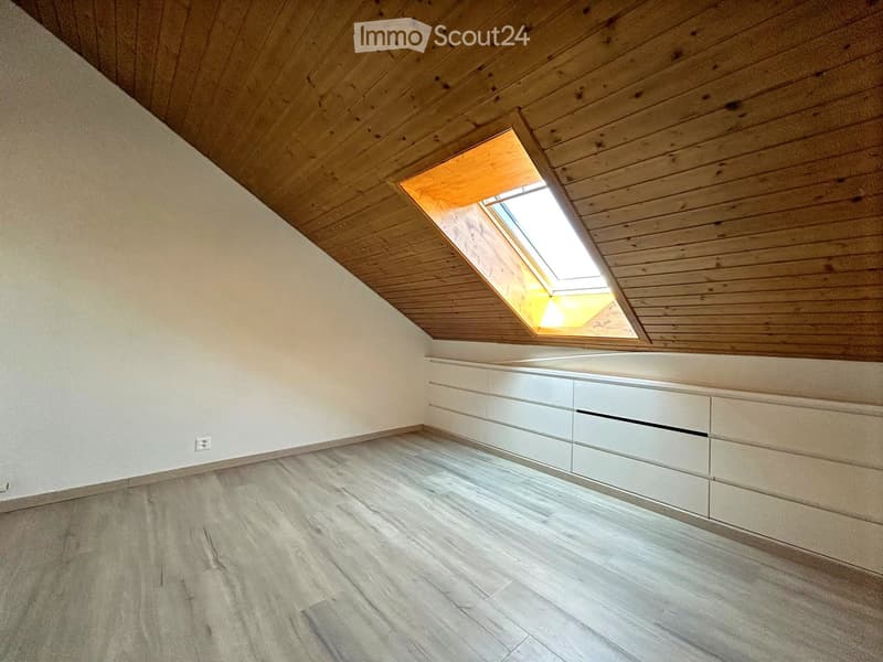 Empty attic room with wooden slatted ceiling, skylight window, built-in white storage units, and light wooden flooring.