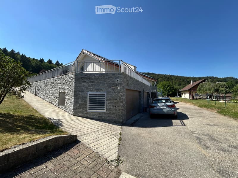 Modern two-story house with stone facade, garage, and raised ground floor leading to a balcony. A car is parked in front of the garage.