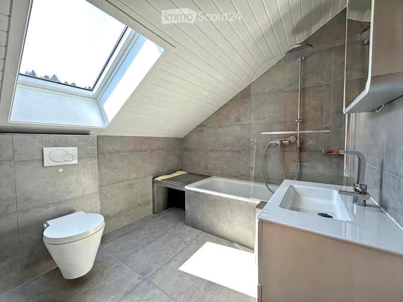 Modern bathroom with grey tiles, white toilet, bathtub, sink, and showerhead. Features a skylight window and a small stool.