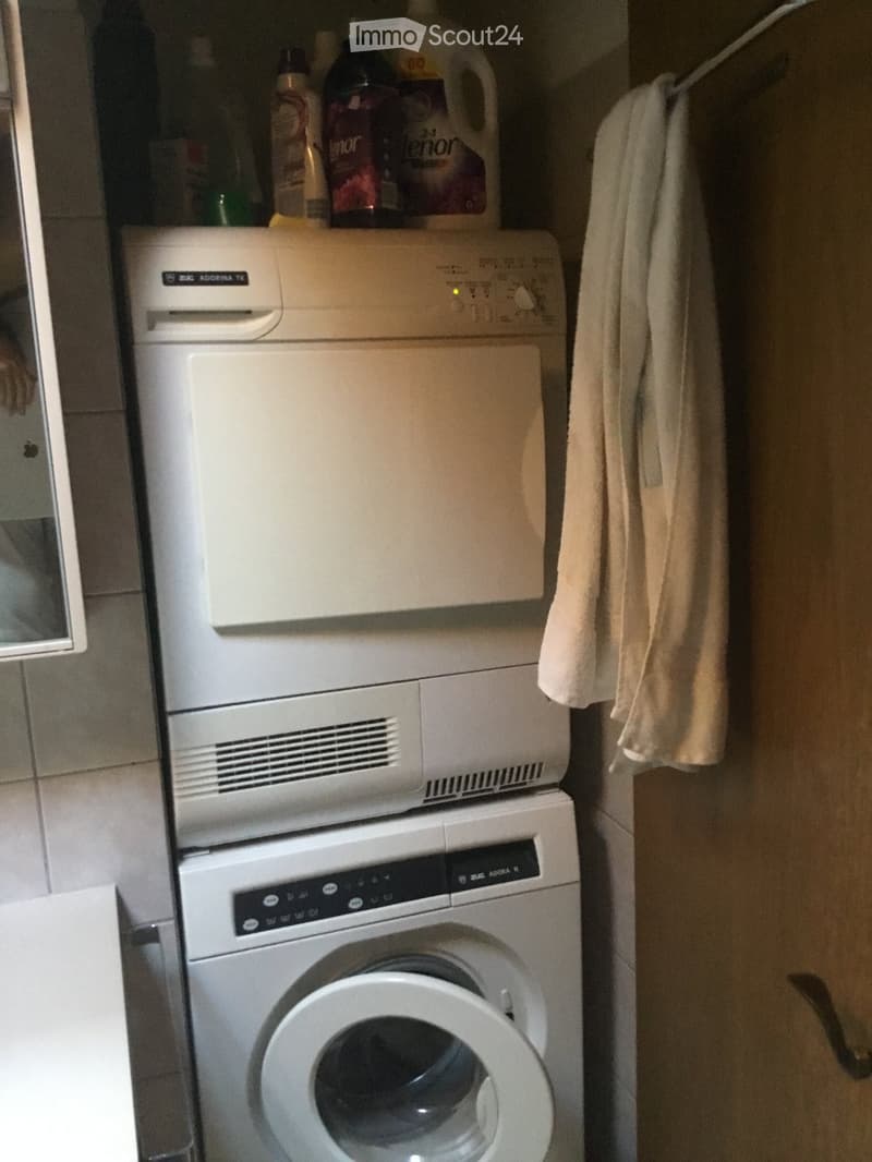A compact bathroom with a stacked washing machine and dishwasher unit, a towel hanging on the side, and cleaning supplies on the shelf above.