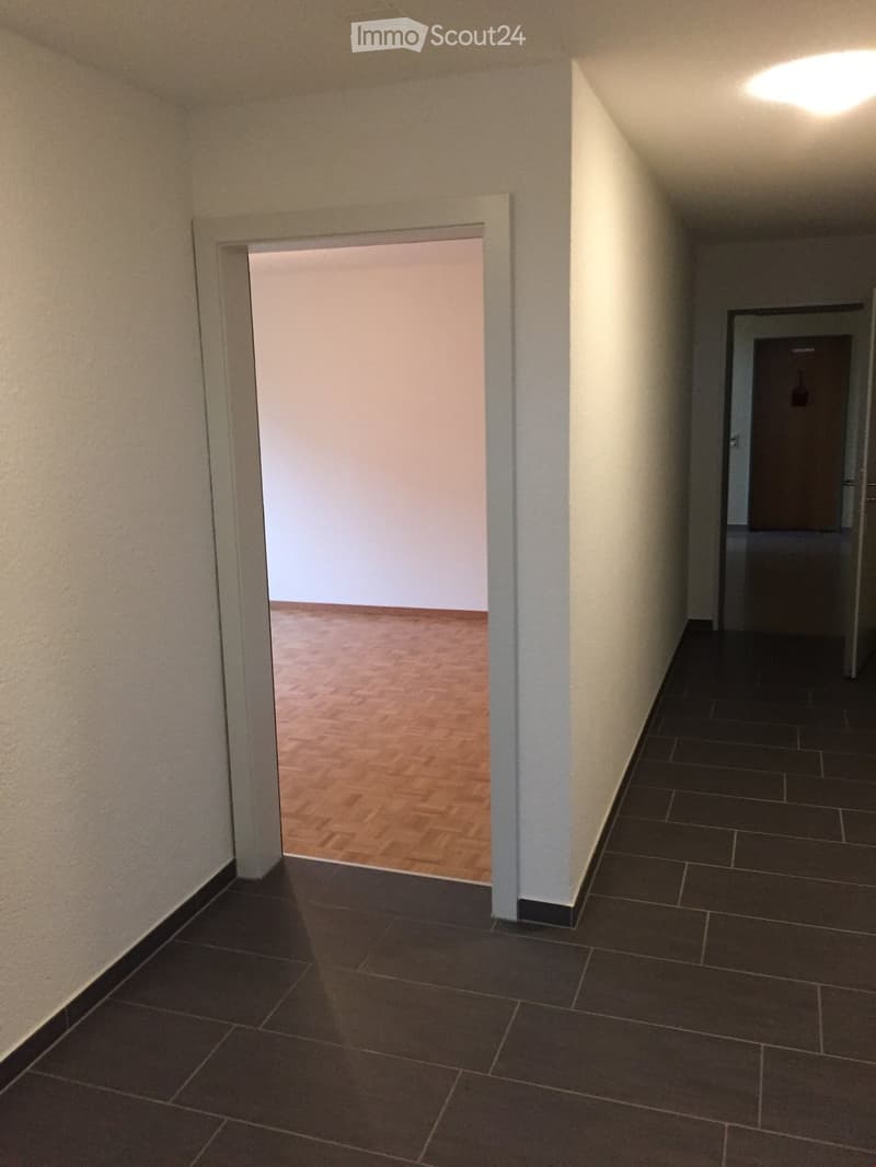 Empty hallway with dark tiled flooring and light-colored walls, featuring an open doorway leading to another room with wooden flooring.