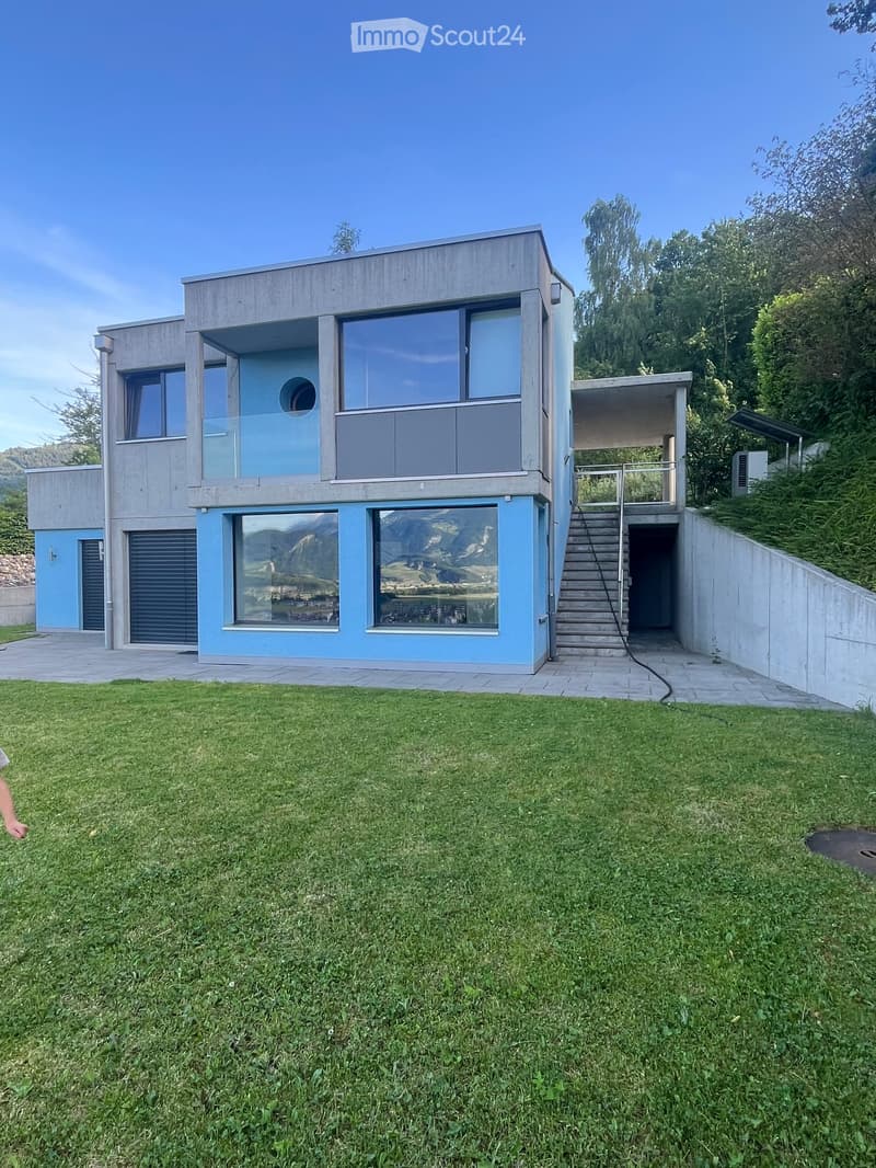 Modern two-story house with light blue and grey exterior, large windows, and a raised ground floor entrance.