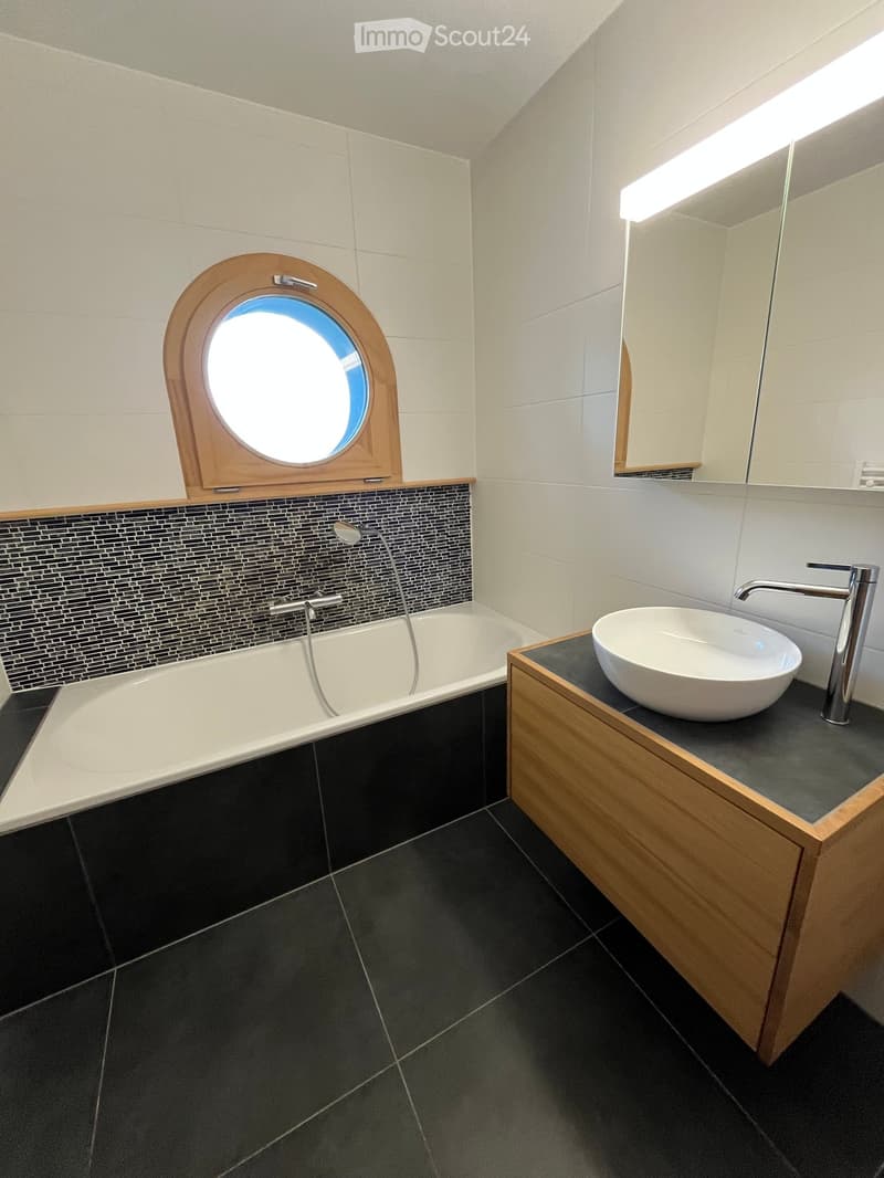 Modern bathroom with a bathtub, sink, and mirror. Features dark tiles, a wooden vanity, and a round window above the bathtub.