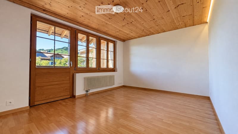 Empty room with wooden flooring, white walls, large windows, and a wooden door leading outside. The room features a radiator and a ceiling light fixture.