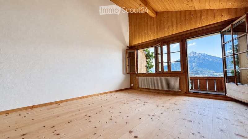 Empty room with wooden flooring, white walls, large windows with wooden frames, and a radiator. The room has a slanted wooden ceiling and offers a scenic mountain view through the windows.