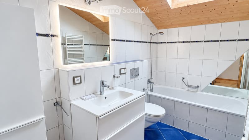 Modern bathroom with white tiles, a sink, bathtub, toilet, and a mirror. Features a wooden slanted ceiling and a radiator.