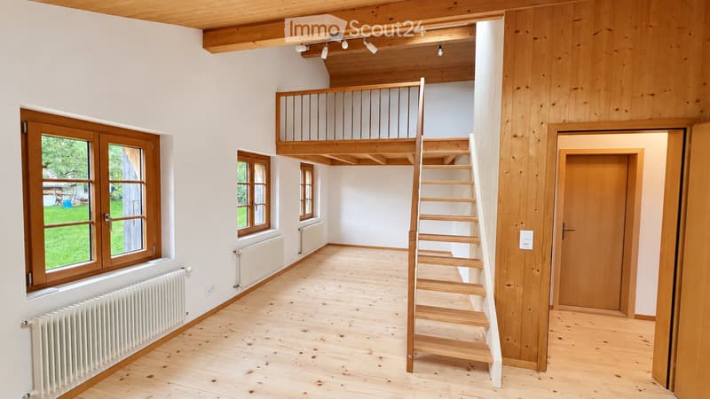 Bright, open room with wooden flooring, white walls, large windows, and a wooden staircase leading to an upper level with a wooden railing.