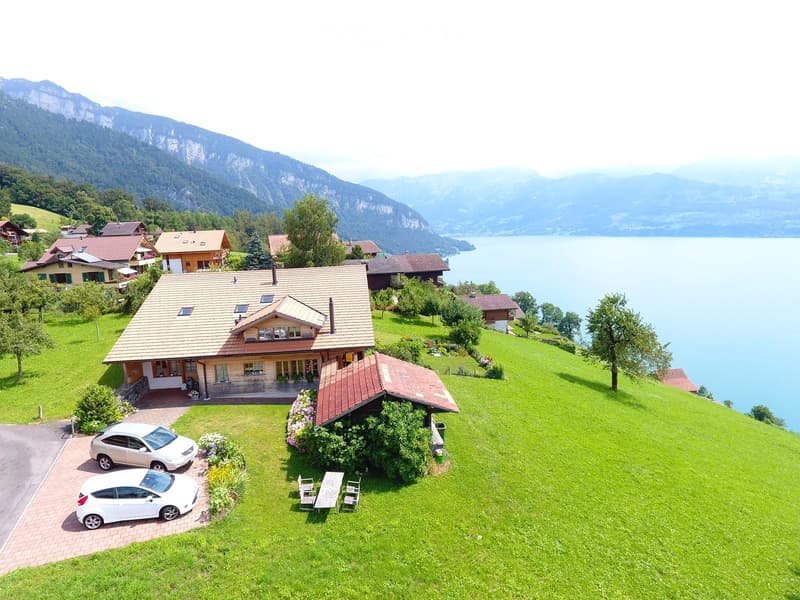 A two-story house with a tiled roof, surrounded by a well-maintained lawn, garden, and a parking area with two white cars. The house is situated near a lake with mountains in the background.