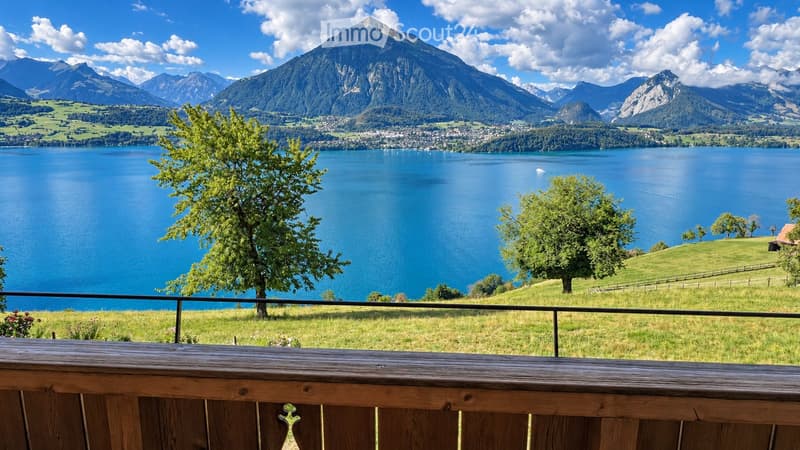 Balcony overlooking a serene lake surrounded by lush green hills and distant snow-capped mountains.