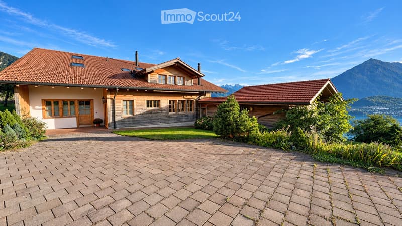 Two-story wooden house with red-tiled roof, large windows, and a paved driveway, surrounded by greenery and a scenic mountain view.