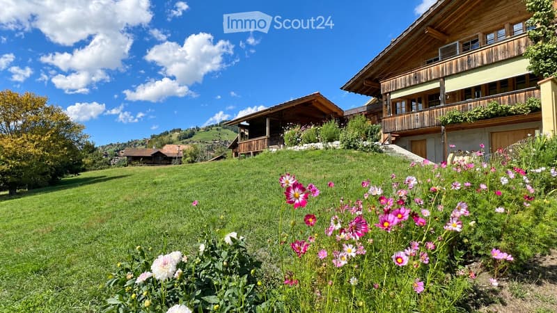 A lush green lawn with colorful flowers in the foreground, surrounded by wooden houses with balconies and a scenic hillside background.
