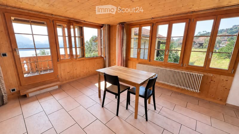 Bright room with wooden walls and ceiling, large windows offering scenic views, tiled floor, and a small dining setup with a table and two chairs.