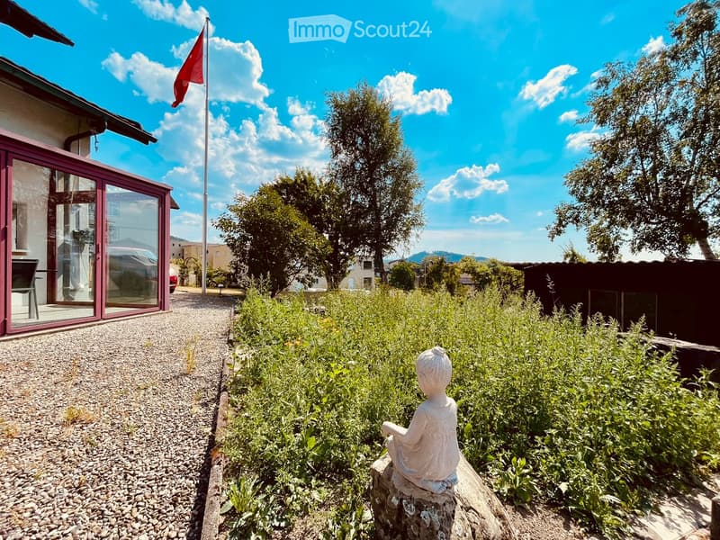 Garden area with gravel pathway, stone statue, lush greenery, and a modern house with large glass doors and a red flagpole.
