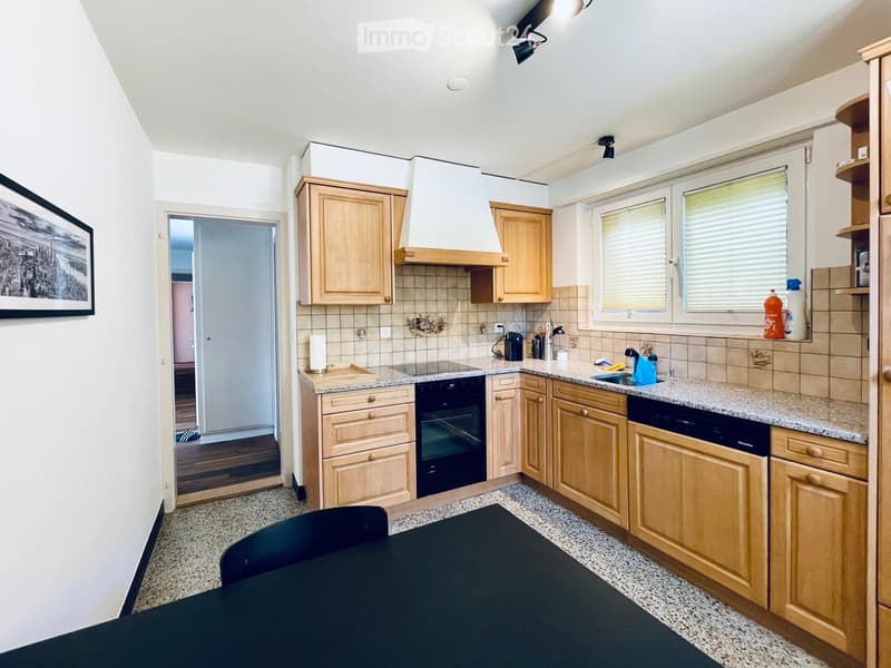 The kitchen features light wooden cabinets, a granite countertop, an oven, a dishwasher, and a small dining table with a black chair. There are windows with blinds and a range hood above the stove.