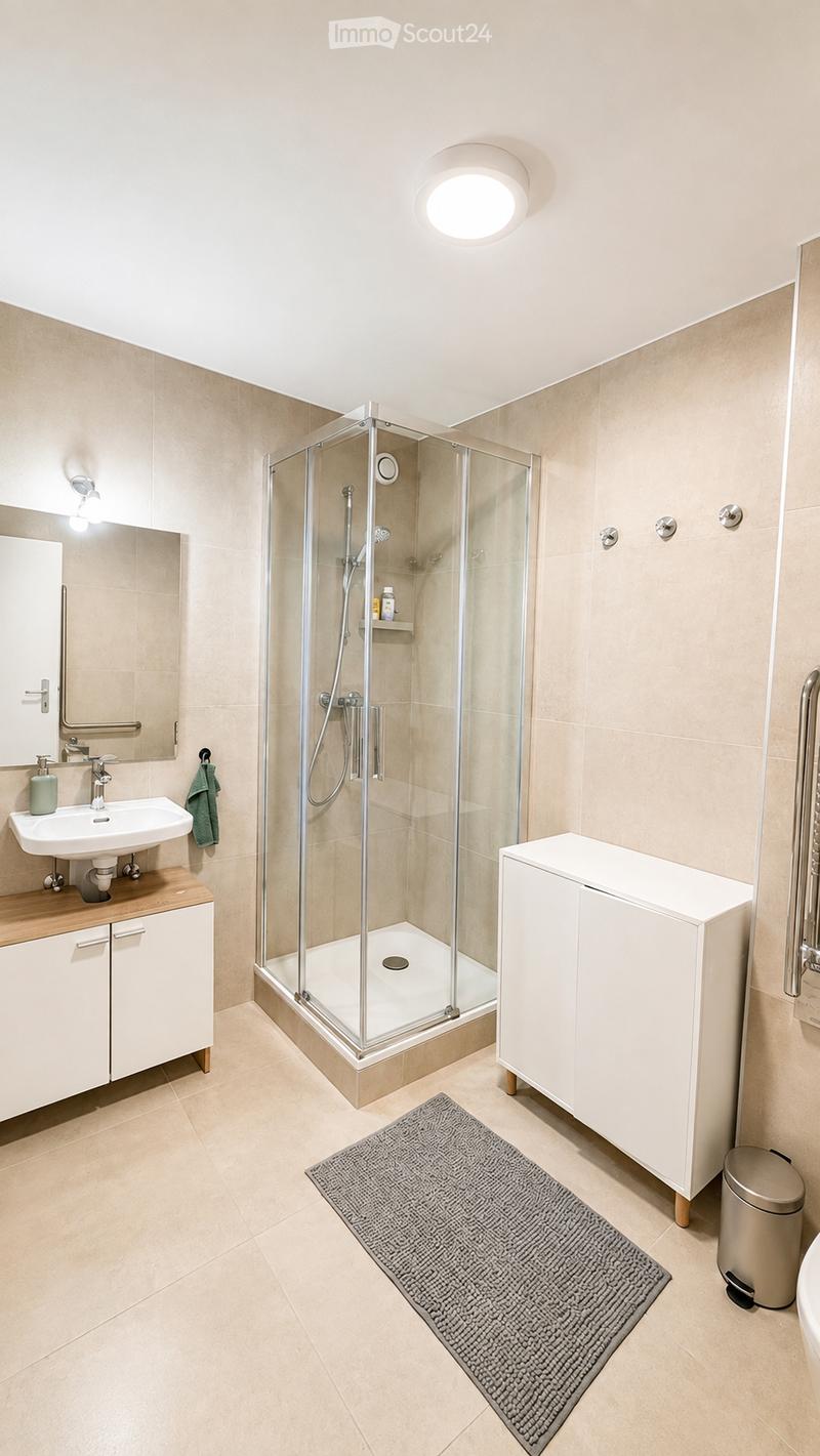 Modern bathroom with beige tiles, a glass-enclosed shower, a white sink with a wooden countertop, a large mirror, and a grey bath mat.