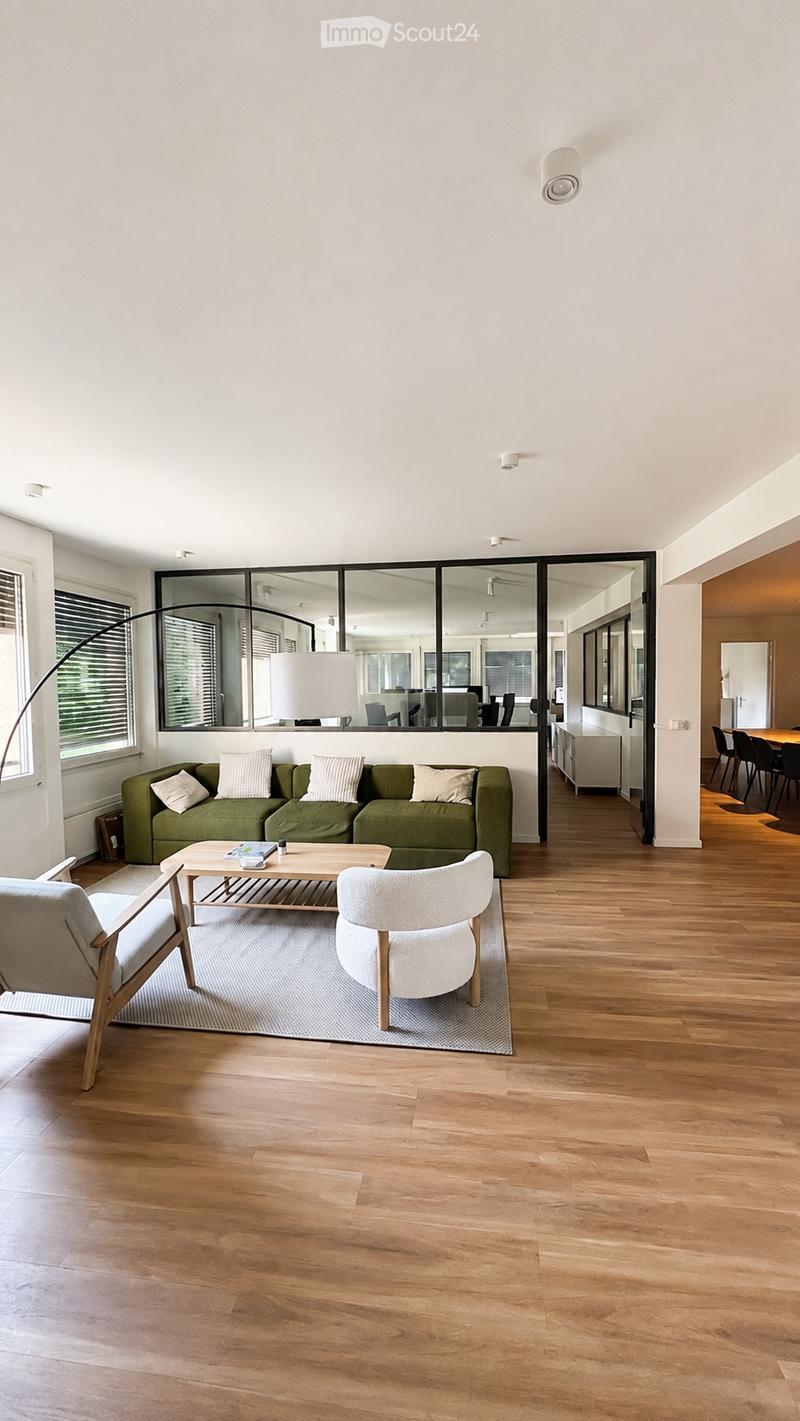 Modern living room with a green sofa, coffee table, two armchairs, floor lamp, and large windows with blinds. The room features wooden flooring and a rug.